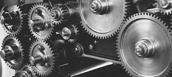 Bachelor Mechatronik Ein Foto von vielen verschiedengroßen Zahnrädern, welche teilweise ineinander greifen. Das Bild ist grau gehalten.