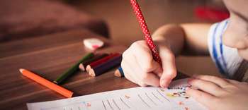 Bachelor Pädagogik Ausschnitt der Hände eines Kindes, welches an einem hölzernen Tisch sitzt. Das Kind malt auf einem Blatt. Verschiedenfarbige Buntstifte liegen neben dem Blatt.