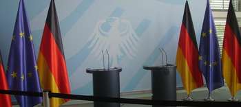 Bachelor Politikwissenschaft Ausschnitt zweier Rednerpulte im Bundestag. Links und rechts davon deutsche sowie europäische Flagge. Inmitten der Rednerpulte der Bundesadler auf hellem Hintergrund.