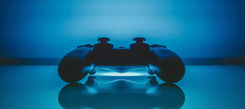 Bachelor Gamedesign Der Controller einer Spielekonsole liegt auf einer gläsernen Platte und spiegelt sich in dieser. Im unteren Zentrum des Bildes ist ein blauer Lichtstrahl erkennbar, welcher das gesamte Bild in eine Blau Farbe taucht.