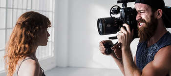 Schauspiel Auf der linken Seite des Bildes befindet sich eine Frau mit langen rötlichen Haaren, auf der rechten Seite des Bildes ein Mann mit Bart, der eine Kappe trägt. Der Mann hält eine professionelle Kamera und scheint die Frau zu filmen. Zu sehen sind die Oberkörper der beiden Personen. Der Raum im Hintergrund ist in einem freundlichen weiß gehalten.