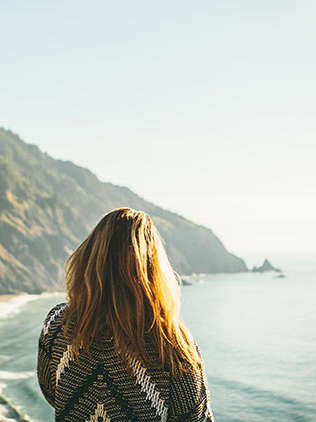Ein Mädchen steht mit dem Rücken zur Kamera. Sie blickt auf einen weiten Strand vor felsiger Landschaft sowie auf das Meer. 