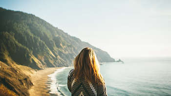 Work & Travel in den USA Ein Mädchen steht mit dem Rücken zur Kamera. Sie blickt auf einen weiten Strand vor felsiger Landschaft sowie auf das Meer.