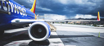 Bachelor Luft- und Raumfahrttechnik Der Ausschnitt eines Flugzeus, welches in blau gehalten ist. Erkennbar ist ein Teil der rechten Seite eines Flugzeugs mitsamt des Triebwerkes. Das Flugzeug befindet sich auf der Startbahn, im Hintergrund ist eine hügelige Landschaft zu sehen.