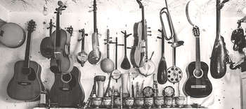 Instrumentalmusik An einer Wand hängen verschiedenste Instrumente durcheinander platziert. Erkennbar sind Gitarren, Trompeten, Trommeln und kleinere Instrumente wie Flöten und exotisch aussehende Instrumente. Das Bild ist in graustufen gehalten.