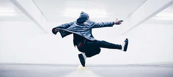 Tanz Im Zentrum des Bildes befindet sich ein Tänzer. Dieser wird in seiner Rückansicht gezeigt und tanzt Breakedance oder HipHop. Er ist sportlich und in blauen Farben gekleidet, trägt eine offene Kapuzenjacke und hat eine Mütze auf. Der Hintergrund ist weiß gehalten.