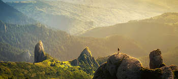 Philosophie Das Bild zeigt eine weite Landschaft. Es ist aus der Luft fotografiert, sodass die unendlich scheinende Landschaft überblickt werden kann. In der Ferne steht eine Person auf einem Felsen und scheint die Weite zu betrachten. Die Landschaft ist geprägt von Hügeln, Wäldern und Felsen, über die sich das Licht der Abendsonne legt.