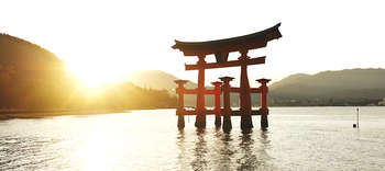 Orientalistik Das Bild zeigt den Itsukushima-Schrein, der seit 1996 zum UNESCO-Weltkulturerbe gehört. Das Tor ist von Wasser umgeben. Im Hintergrund ist eine bergige Landschaft erkennbar, durch die Sonnenstrahlen scheint.