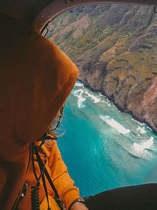 Eine Person sitzt in in einem Hubschrauber und schaut aus dem Fenster. In diesem ist eine bergige Landschaft und ein türkisblaues Meer zu sehen. 