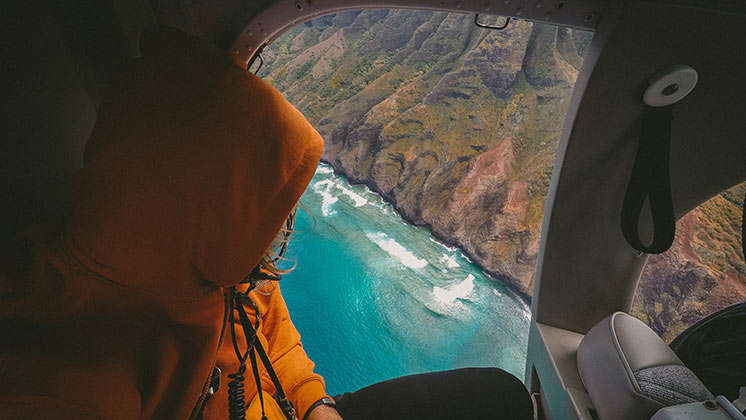 Werde zum Traveller Eine Person sitzt in in einem Hubschrauber und schaut aus dem Fenster. In diesem ist eine bergige Landschaft und ein türkisblaues Meer zu sehen.