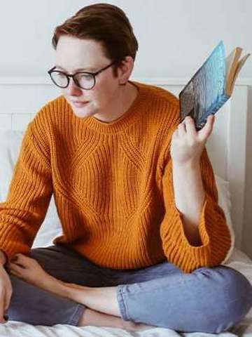 Ein Mädchen sitzt mit ihrem Laptop im Schneidersitz auf ihrem Bett und hält ein Buch in der linken Hand.