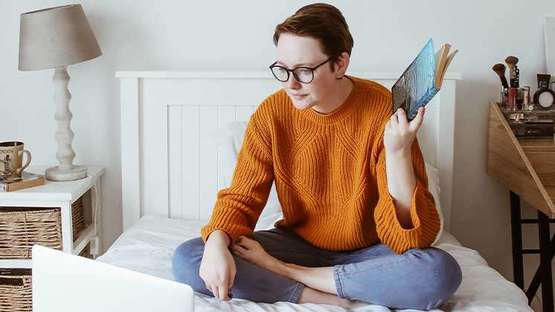 Ein Mädchen sitzt mit ihrem Laptop im Schneidersitz auf ihrem Bett und hält ein Buch in der linken Hand.