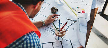 Bachelor Bauingenieurwesen Ein Bauchzeichner bei der Arbeit. Sichtbar ist der Rücken des Mannes, der eine orangene Weste trägt und auf einem Bauplan zeichnet.