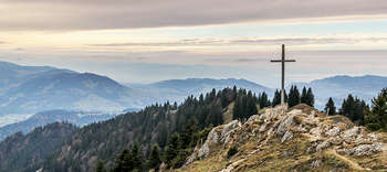 Religionswissenschaften Das Bild zeigt einen Gipfel, von dem aus die weite Landschaft überblickt werden kann. Auf dem Gipfel befindet sich ein hölzernes Gipfelkreuz.