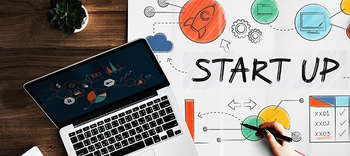 Bachelor BWL Foto eines Holztisches, auf dem ein Laptop steht und ein großes Plakat liegt. Dazu die Hand einer Person, die etwas auf das Plakat schreibt. Das Plakat prägt mehrere Icons sowie das Wort START UP in der Mitte.