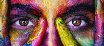 Kunst Das Bild zeigt die Nahaufnahme eines Gesichtes von den Augenbrauen bis zur Nase. Im Fokus des Bildes stehen die ausdrucksstarken braunen Augen der Person. Das Gesicht ist kunstvoll mit zahlreichen bunten Farben bemalt, sodass die Haut nicht mehr erkennbar ist. Die Person legt dabei ihre Hände um ihr Gesicht, welche ebenfalls künstlerisch bemalt sind.
