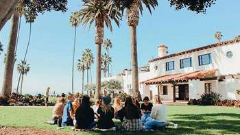 Sprachkurse im Ausland Eine Gruppe Jugendlicher sitzt in einem Kreis auf einem gepflegten Rasen vor einem weißen Haus in mediterranen Stil. Auf dem Grundstück viele Palmen und weiteren Gewächsen.