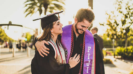 Ein Mann und eine Frau in Graduations-Kleidung und mit Akademischen Hüten vor einem sommerlichen Hintergrund aus Palmen. 