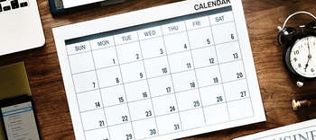 Bachelor Facility Management Ausschnitt eines Holztisches, auf dem ein Kalender liegt. Links daneben deutet sich ein Handy an, rechts eine Uhr.