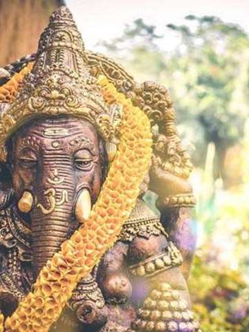 Abbildung einer Statue der Hindu-Gottheit Ganesha. Im Hintergrund eine Kulisse aus Pflanzen und Palmen durch die Sonnenstrahlen scheinen.