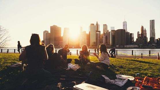 Eine Gruppe junger Menschen sitzt in der Abenddämmerung an einem See. Im Hintergrund ist die Skyline einer Großstadt zu sehen.