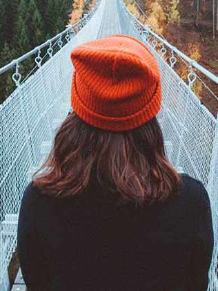Ein Mädchen mit einer orangenen Pudelmütze steht mit dem Rücken zum Betrachter am Rand einer Hängebrücke. Es ist Herbst.