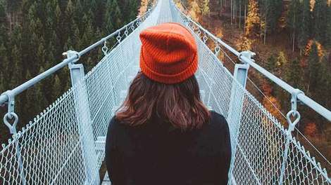 Ein Mädchen mit einer orangenen Pudelmütze steht mit dem Rücken zum Betrachter am Rand einer Hängebrücke. Es ist Herbst.