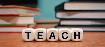 Bachelor Lehramt Nahaufnahme eines Schreibtisches. Im Hintergrund sind verschwommen zwei Bücherstapel erkennbar. Im Fokus des Bildes fünf Buchstabenwürfel, die das Wort TEACH bilden.