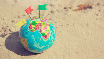 Sprachreisen Eine Weltkugel an einem Sandstrand. In die Kugel sind drei kleine bunte Fahnen gesteckt, welche Orte markieren.