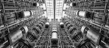 Bachelor Verfahrenstechnik Das Bild zeigt das Innere einer Fabrik aus der Froschperspektive. Das Innere des Gebäudes besteht aus vielen gestellartigen Etagen, die durch einen riesigen Innenhof miteinander verknüpft scheinen. Das Bild ist in graustufen gehalten und scheint stählern und metallisch.