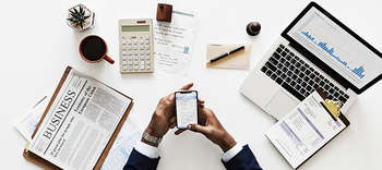 Bachelor Accounting Heller und moderner Schreibtisch, auf dem ein Laptop, eine Business-Taschenrechnung, ein Taschenrechner und ein Klemmbrett liegt. An dem Tisch sitzt ein Mann, der eine silberne Uhr und einen Anzug trägt. Er tippt etwas auf seinem Handy.