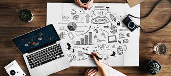 Bachelor VWL Ausschnitt eines Schreibtisch, an dem zwei Personen arbeiten. Darauf ein Laptop sowie ein Plakat mit gezeichneten Symbolen aus dem Wirtschaftsbereich.