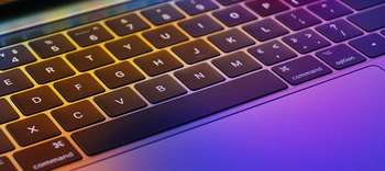 Computerlinguistik Eine Nahaufnahme von einer glatten Laptop-Tastatur. Die Tastatur wird im oberen Bereich leicht gelblich angestrahlt und im unteren Bereich violett.