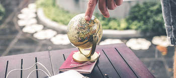 Bachelor IBMS Ein Gartentisch, auf dem ein goldenener kleiner Globus steht. Eine Person hält ihren Finger auf diesem. Im Hintergrund lässt sich eine Gartenanlage erahnen.