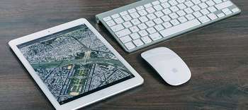 Geoinformatik Auf einer Arbeitsplatte aus dunklem Holz liegen ein Tablet, eine Tastatur und eine kabellose Computermaus. Die Gegenstände sind gerade angeordnet und haben alle eine weiße Farbe.