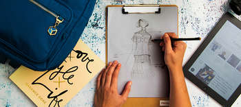 Fashion Design Auf einem Schreibtisch liegt ein Klemmbrett mit einem Papier, auf das eine Person zeichnet. Der Fokus der Zeichnung liegt auf der Kleidung, welche designt wird. Rechts des Klemmbrettes liegt ein Table. Dieses zeigt einen einen Artikel mit dem Schlagwort: FASHION. Links des Klemmbrettes mit der Zeichnung liegt ein blauer Rucksack.