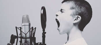 Gesang Das Bild zeigt Kopf und Hals eines Jungen, welcher in ein Studio-Mikrofon sing. Er hat die Augen zum Singen geschlossen und den Mund weit aufgerissen. Das Bild ist in graustufen gehalten.