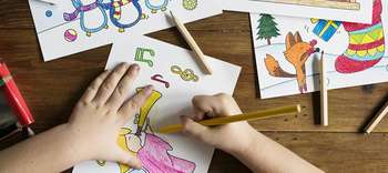 Bachelor Sozialpädagogik Ausschnitt eines Holztisches, auf dem verschiedene Ausmalbilder und Stifte liegen. Zu sehen sind die Hände eines Kindes, die eines der Bilder mit Buntstiften ausmalen.