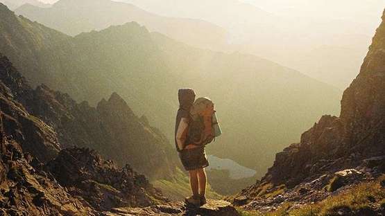 Eine Person steht in einer bergigen Landschaft, durch die einige Sonnenstrahlen fallen. Sie trägt einen Rucksack.