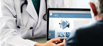 Bachelor Medizin Ausschnitt zweier Personen, die sich gegenüber sitzen. Die Person, welche von vorne zu sehen ist, trägt einen Arztkittel und ihr Hals schmückt ein Stethoskop. Die Person zeigt der gegenübersitzenden Person etwas auf einem Tablet.