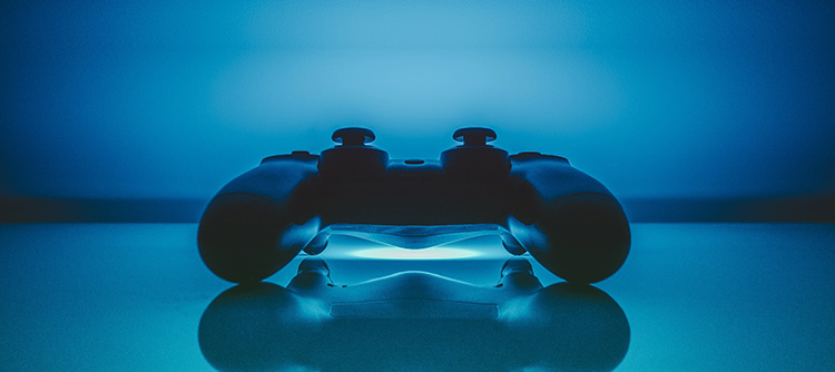Bachelor Gamedesign Der Controller einer Spielekonsole liegt auf einer gläsernen Platte und spiegelt sich in dieser. Im unteren Zentrum des Bildes ist ein blauer Lichtstrahl erkennbar, welcher das gesamte Bild in eine Blau Farbe taucht.