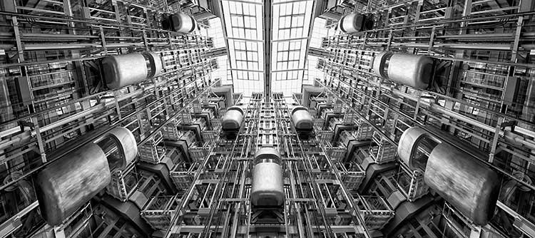 Bachelor Verfahrenstechnik Das Bild zeigt das Innere einer Fabrik aus der Froschperspektive. Das Innere des Gebäudes besteht aus vielen gestellartigen Etagen, die durch einen riesigen Innenhof miteinander verknüpft scheinen. Das Bild ist in graustufen gehalten und scheint stählern und metallisch.