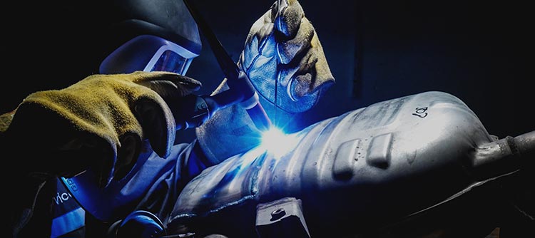 Bachelor Maschinenbau Das Bild zeigt den Ausschnitt einer Person, welche die Tätigkeit des Schweißens ausführt. Die Person trägt eine Schutzbrille sowie gelbe Handschuhe. Durch das Schweißverfahren ist ein blauer heller Lichtpunkt erkennbar.