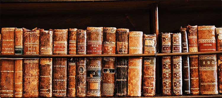 Theologie Eine Nahaufnahme von einem Bücherregal. In dem Regal steht eine vielzahl antiker Bücher aneinander gereiht. Diese sind mit schmuckvollen goldenen Elementen und Mustern verziert. Die Farben wirken rustikal, braun, rot und orange dominieren.