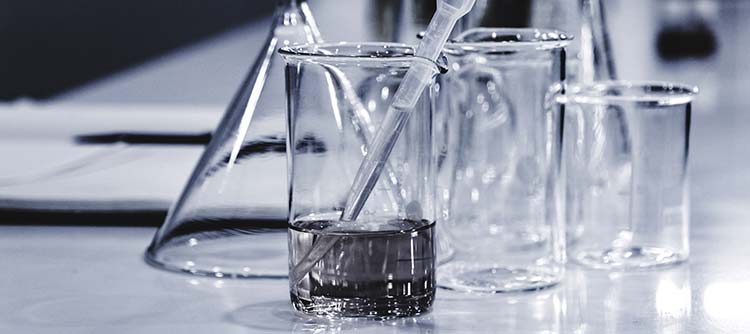 Bachelor Chemie Nahaufnahme eines Tisches in einem Chemielabor. Auf dem Tisch stehen drei Bechergläser in verschiedenen Größen. Das Bächerglas in der Mitte enthält eine Flüssigkeit. Im Hintergrund deuten sich weitere Kolben und Gefäße an. Das Bild ist in graustufen gehalten.