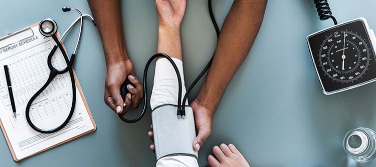 Bachelor Gesundheitswissenschaften Ausschnitt des Schreibtisches eines Arztes. Patient und Arzt sitzen sich gegenüber. Der Arzt misst den Blutdruck des Patienten.