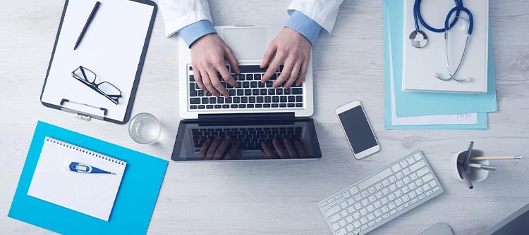 Bachelor Pflegemanagement Ausschnitt des Schreibtisches eines Arztes. Auf dem Schreibtisch ein Laptop, Handy, Klemmbrett, Notizblock sowie ein Stethoskop. Eine Person tippt am Laptop. Sie trägt einen Ärztekittel.