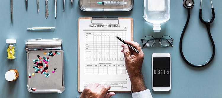 Bachelor Pflegewissenschaften Ausschnitt des Schreibtisches eines Arztes. Verschiedene medizinische Instumente sind auf diesem symmetrisch angeordnet. Darunter eine Brille, Klemmbrett, Stethoskop und Medikamente. Zu sehen sind auch die Hände des Arztes, der etwas auf dem Klemmbrett notiert.