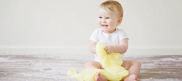 Bachelor Early Years Studies Ein Baby sitzt auf einem hölzernen Fußboden und hält ein gelbes Tuch in der Hand. Das Baby trägt einen hellen body und lacht.