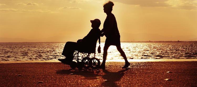 Bachelor Sonderpädagogik Bild von einem Strand im Sonnenuntergang. Im Zentrum des Bildes die Konturen zweier Personen. Die eine Person schiebt die andere in einem Rollstuhl an dem Strand entlang.